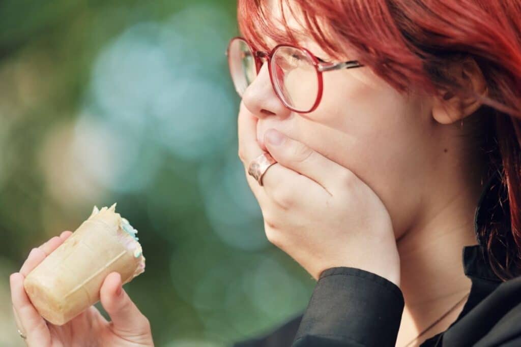 Woman with red hair and glasses eating a sandwich, experiencing tooth sensitivity.
