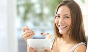 Woman smiling while eating cereal with berries from a bowl during dental recovery.