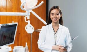 Smiling female dentist in white coat holding tablet in modern dental office with surgical light.