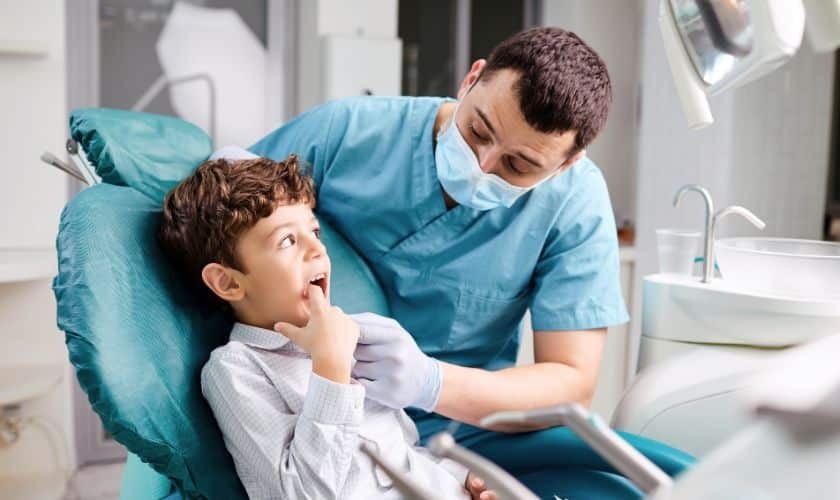Young boy smiling in dental chair while pediatric dentist examines his teeth during first visit.