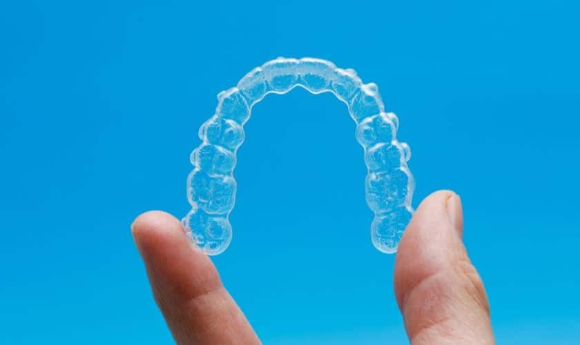 Clear Invisalign aligner tray held between fingers against blue background.