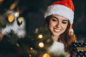 Smiling woman in Santa hat and white sweater with holiday lights and decorated tree in background.