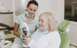 Dental patient admiring her smile in mirror while hygienist smiles nearby in treatment room.