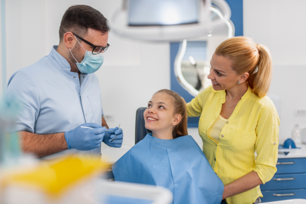 Dentist showing dental tools to young patient while mother smiles beside her in treatment room.