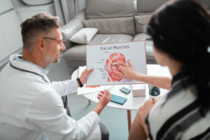 Male dentist explaining facial muscles diagram to female patient in consultation.