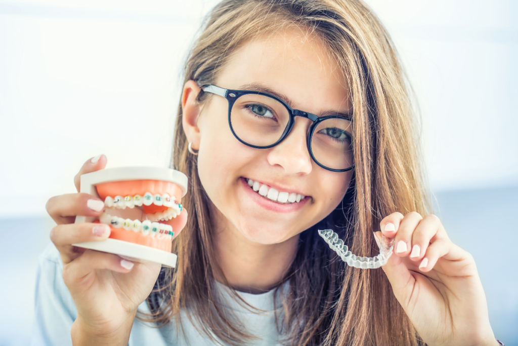 Smiling girl with glasses holding dental model with braces and clear aligner tray.